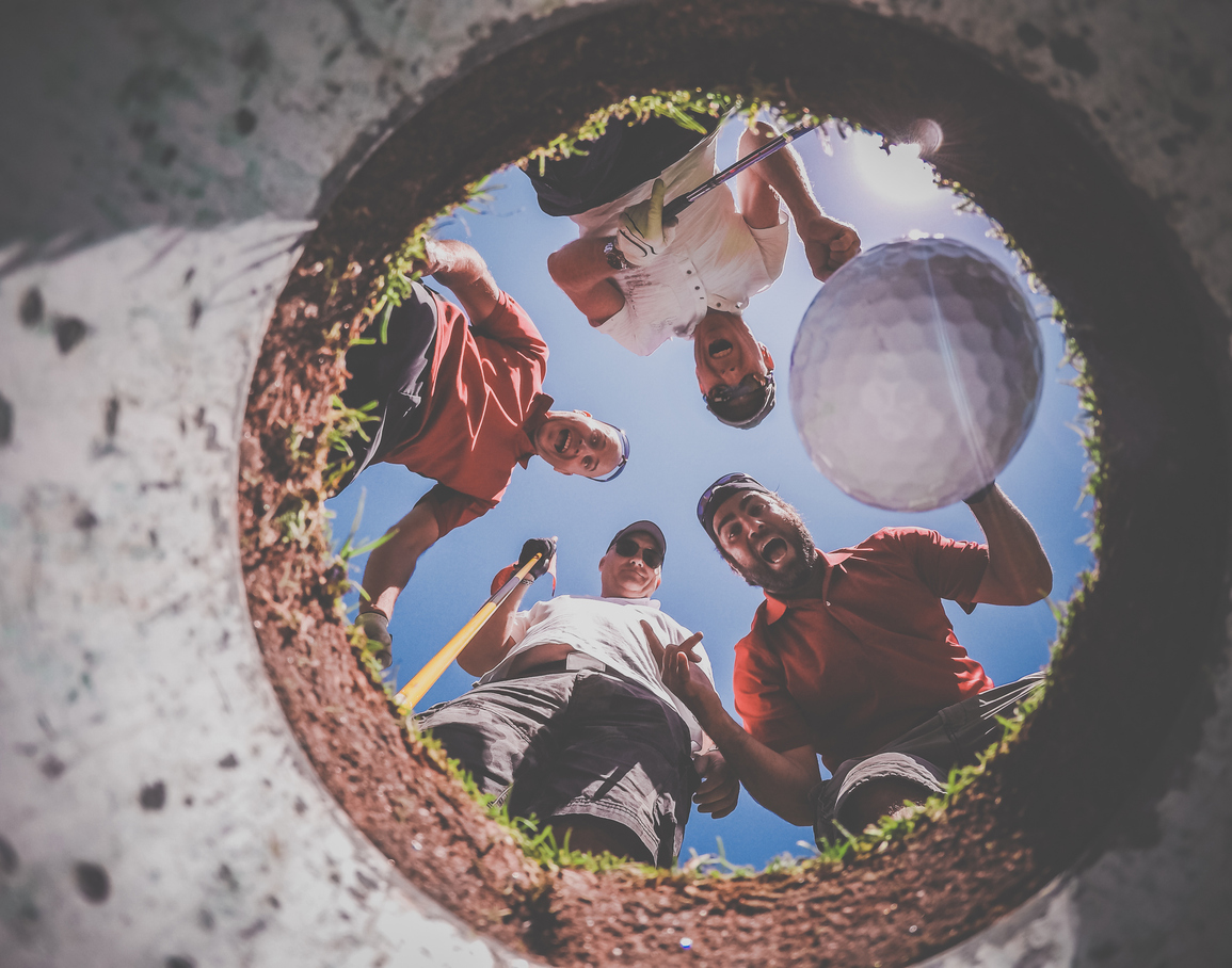 A Point Of View picture of 4 Golf Players and Golf Ball view From Inside the Hole on a sunny day of summer. The players are excited and happy.